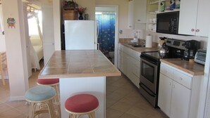 A compact kitchen with a central island, a stove, microwave, and basic appliances. There are stools at the island and a small dining area with a chair. - Sunrises & Sunsets Tropical Getaway - Incredible Views Overlooking Caribbean Sea (St. Thomas)