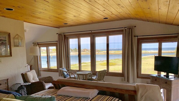 A room with a bed, a TV, a desk, and large windows offering a view of the sea.