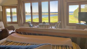 A room with a bed, a table, and chairs, large windows with a view of the sea and beach.