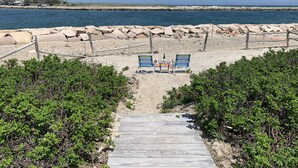 A beach with two blue lounge chairs and a bottle on a small table, a wooden pathway leading to the beach, and a rocky breakwater in the background.