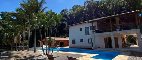 Una terraza de madera con sillones, una piscina y un edificio con balcón.