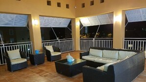 Un espacio de descanso al aire libre con mobiliario de mimbre, incluyendo sofás y sillones, y piso de baldosas.