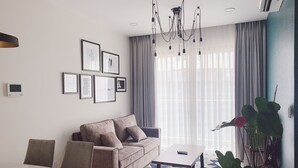 Ein modernes Wohnzimmer mit einer Couch, einem Couchtisch und einem Essbereich. An der Wand hängen gerahmte Bilder, es gibt eine Kristallleuchter und ein großes Fenster mit Vorhängen.