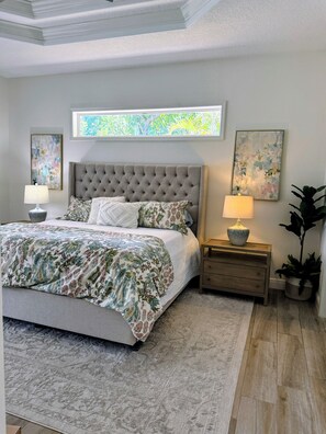 Une chambre à coucher avec une tête de lit capitonnée, une couvre-lit à motifs floraux, deux tables de chevet avec des lampes, une grande fenêtre offrant une belle vue et une plante en pot.