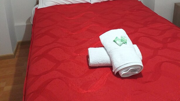 A single bed with a red quilt and white pillows, a folded white towel with a green tag, and a painting of flowers on the wall. - Filfor Hause (Cusco)