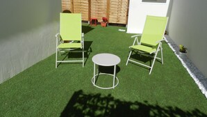 Un pequeño patio con dos sillas verdes y una mesa redonda, rodeado de césped artificial.