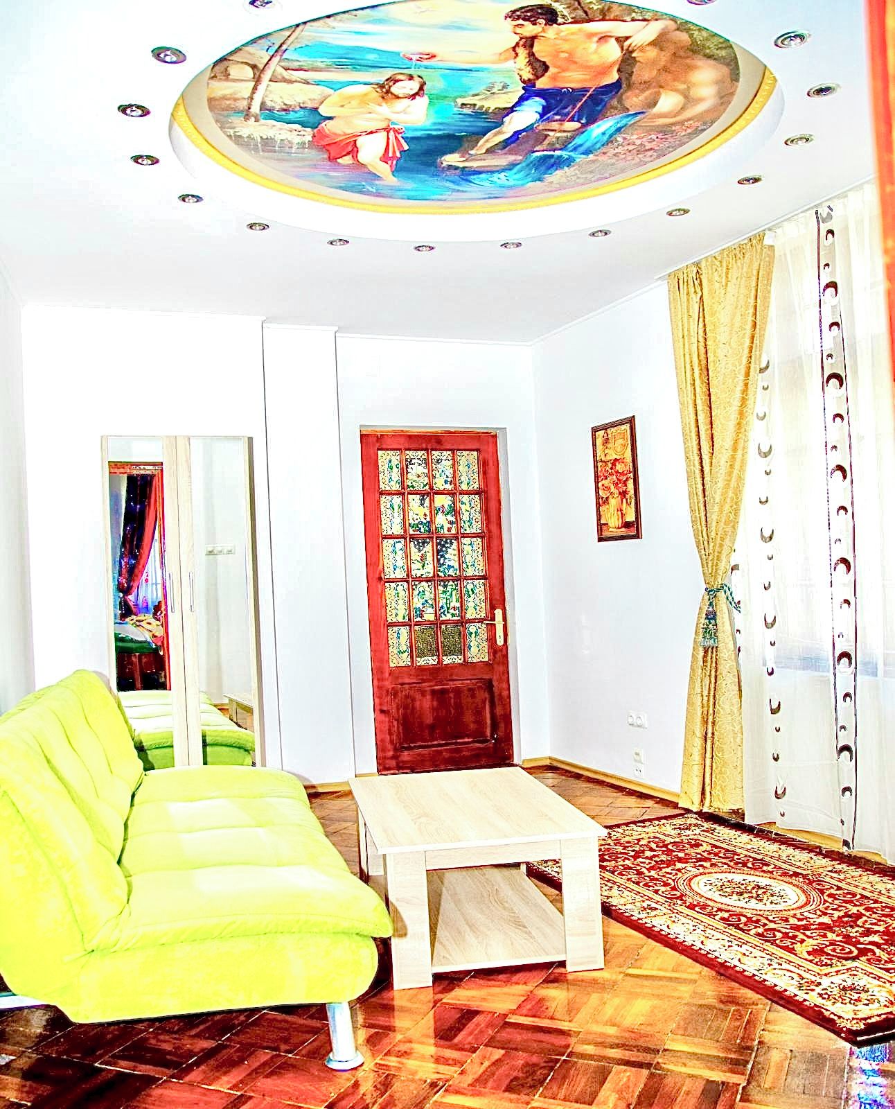 Une pièce avec un plancher en bois, un canapé vert, une petite table, une porte rouge ornée d’un panneau décoratif et une fresque murale au plafond.
