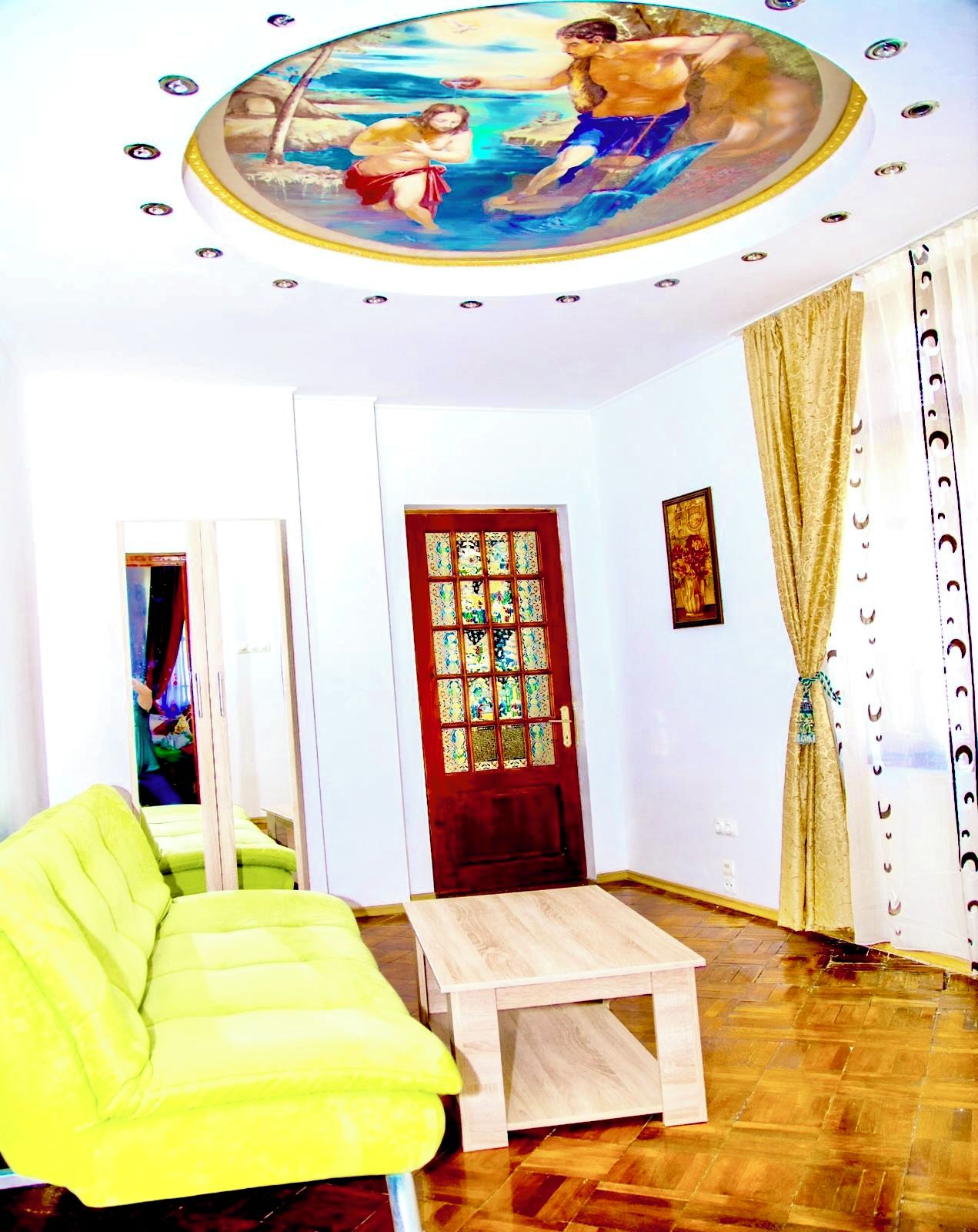 A room with a wooden floor, a green sofa, a wooden coffee table, a red door with decorative glass panels, and a ceiling mural.