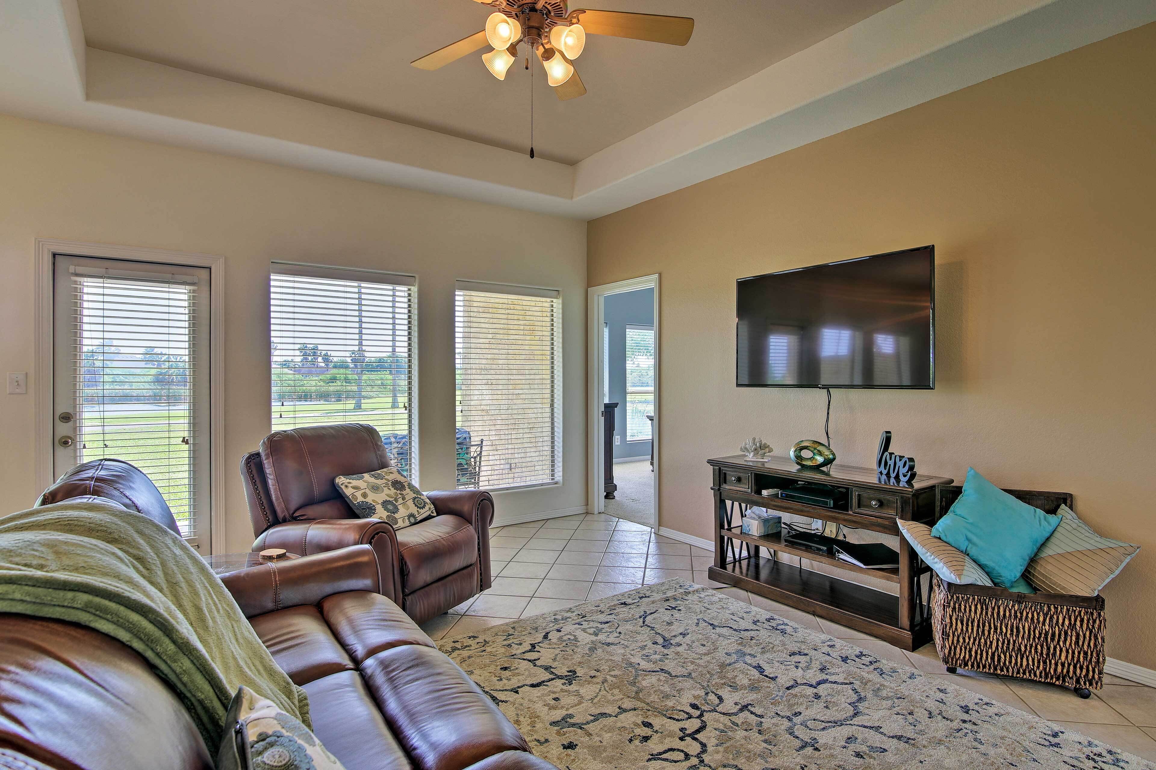 A spacious living room with a leather sofa, a coffee table, and a flat-screen TV mounted on the wall. There are large windows with blinds, a ceiling fan, and a patterned rug on the floor.