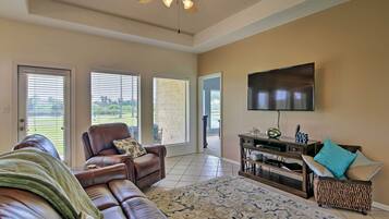 A spacious living room with a leather sofa, a coffee table, and a flat-screen TV mounted on the wall. There are large windows with blinds, a ceiling fan, and a patterned rug on the floor.