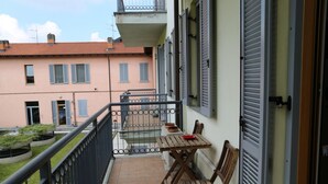A balcony with a wooden table and chairs, a small garden area, and a building with shutters in the background.