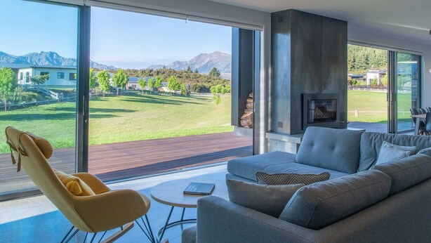 A modern living room with a large sliding glass door, a grey sofa, a yellow rocking chair, and a fireplace. - The Gem in Peninsula Bay (Wanaka)