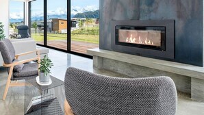 A modern living room with a fireplace, a sofa, and a view of the outdoors. - The Gem in Peninsula Bay (Wanaka)