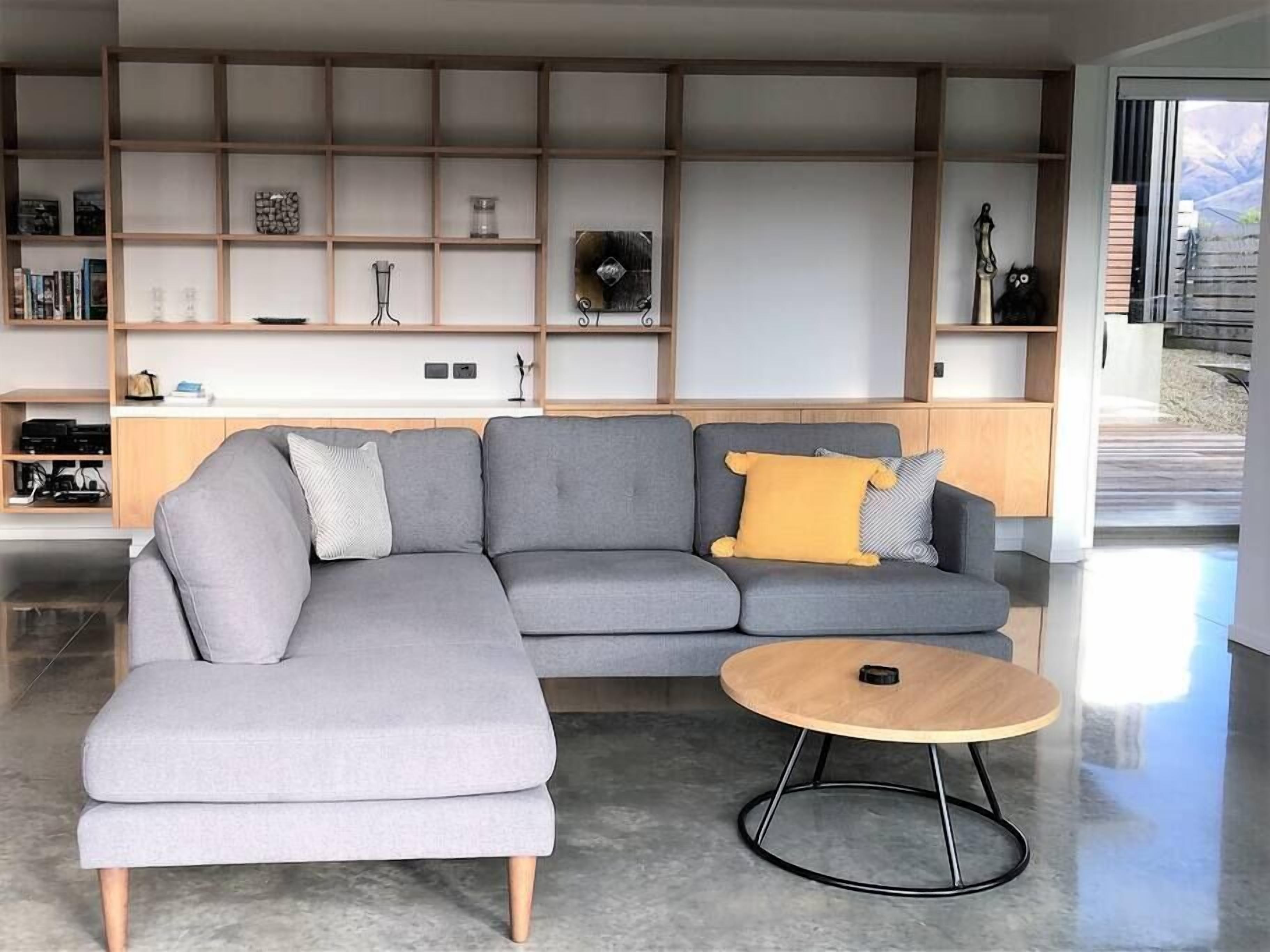 Een moderne woonkamer met een grijze hoekbank, een klein rond houten bijzettafeltje en een inbouwplank met diverse decoratieve objecten.