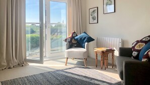 A room with a sliding glass door leading to a balcony, a white armchair with a colorful cushion, a dark sofa, a patterned rug, and two framed botanical prints on the wall. - ‘Waterside’ is a 1st floor apartment  on the river in Rye, East Sussex (Rye, East Sussex)
