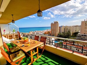 Een balkon met een houten tafel en stoelen, uitzicht op de zee, en gebouwen op de achtergrond.