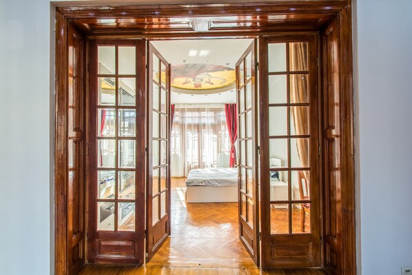 A hallway with wooden doors leading to a bedroom with a bed and a desk.