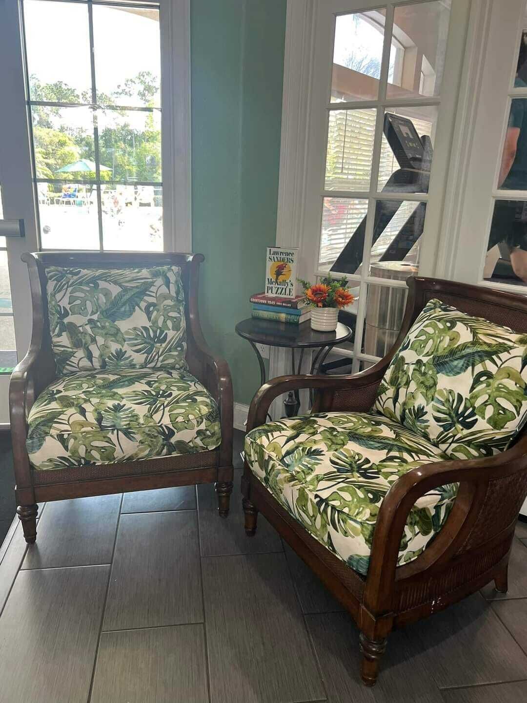 To læderarmstole med tropisk blad-betræk, et lille bord med en bog og blomster, og udsigt til en parkeringsplads gennem store vinduer.