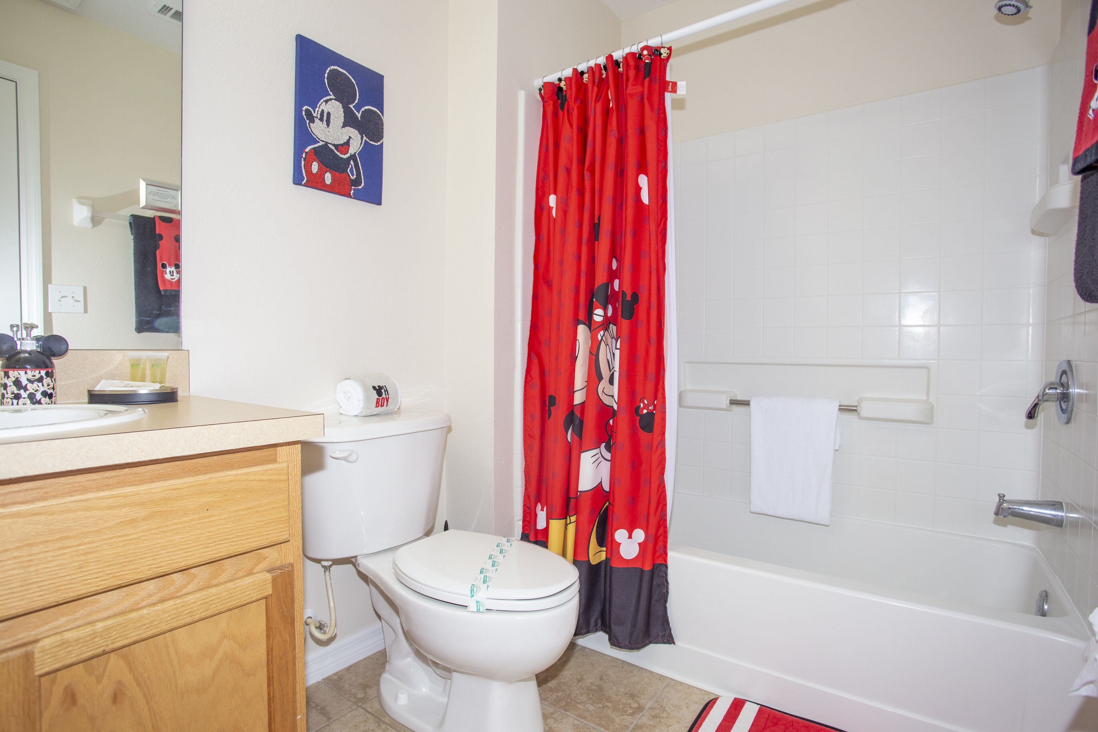 A bathroom with a Mickey Mouse themed shower curtain, a white toilet, a wooden vanity, and a bathtub.