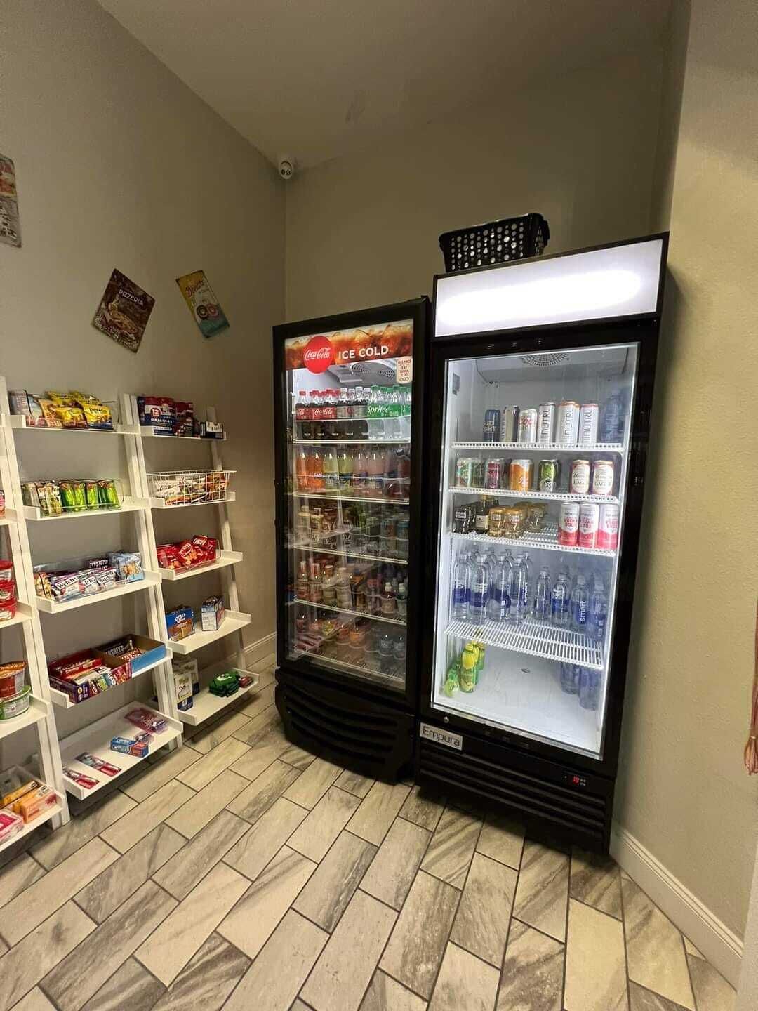 Rincón con dos dispensadores de bebidas, uno con el rótulo de Coca-Cola y otro con el de Sprite, y estantes llenos de aperitivos y bebidas.