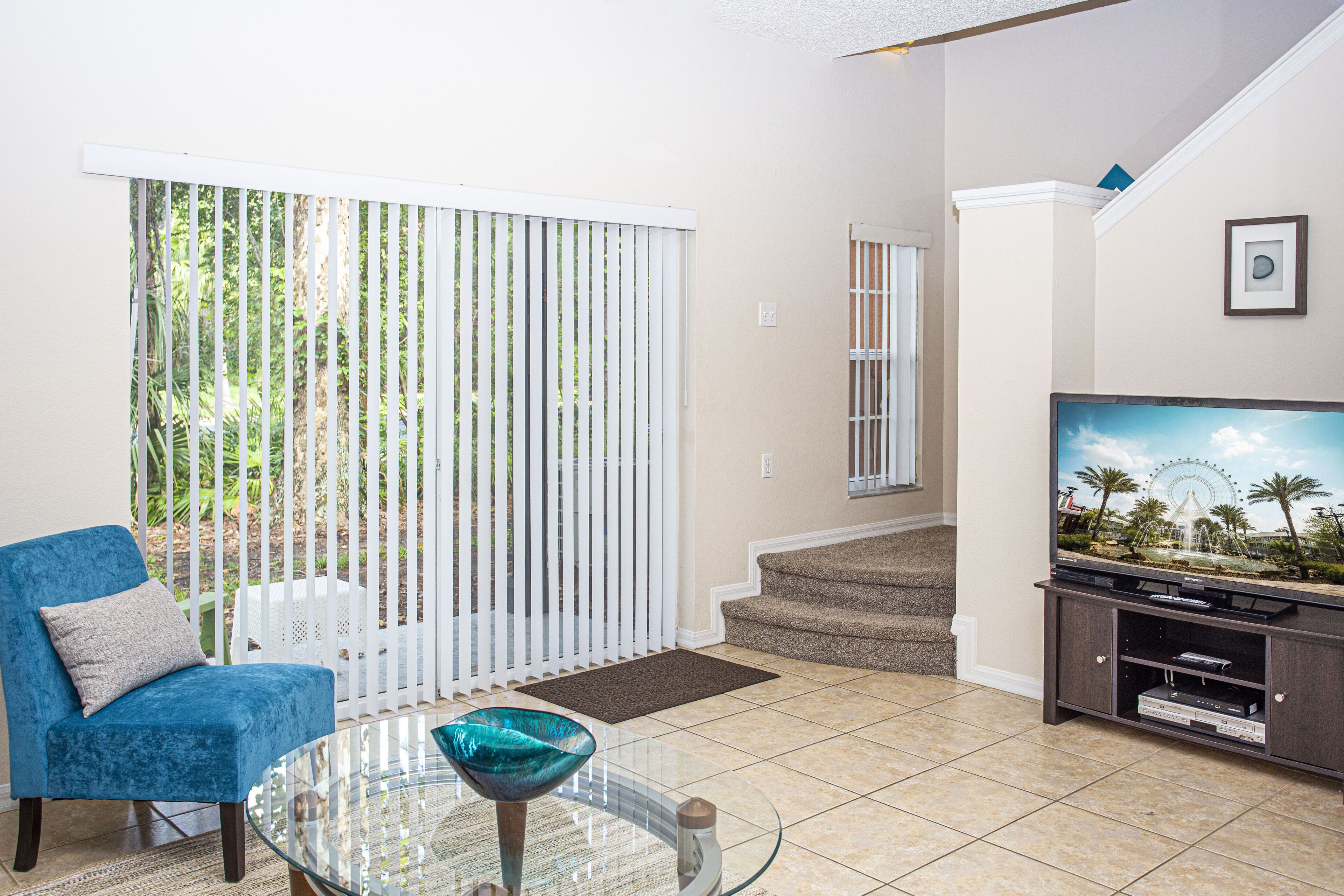 A living room with a blue chair, a glass table, a TV on a stand, and a view of a white fence and greenery outside.