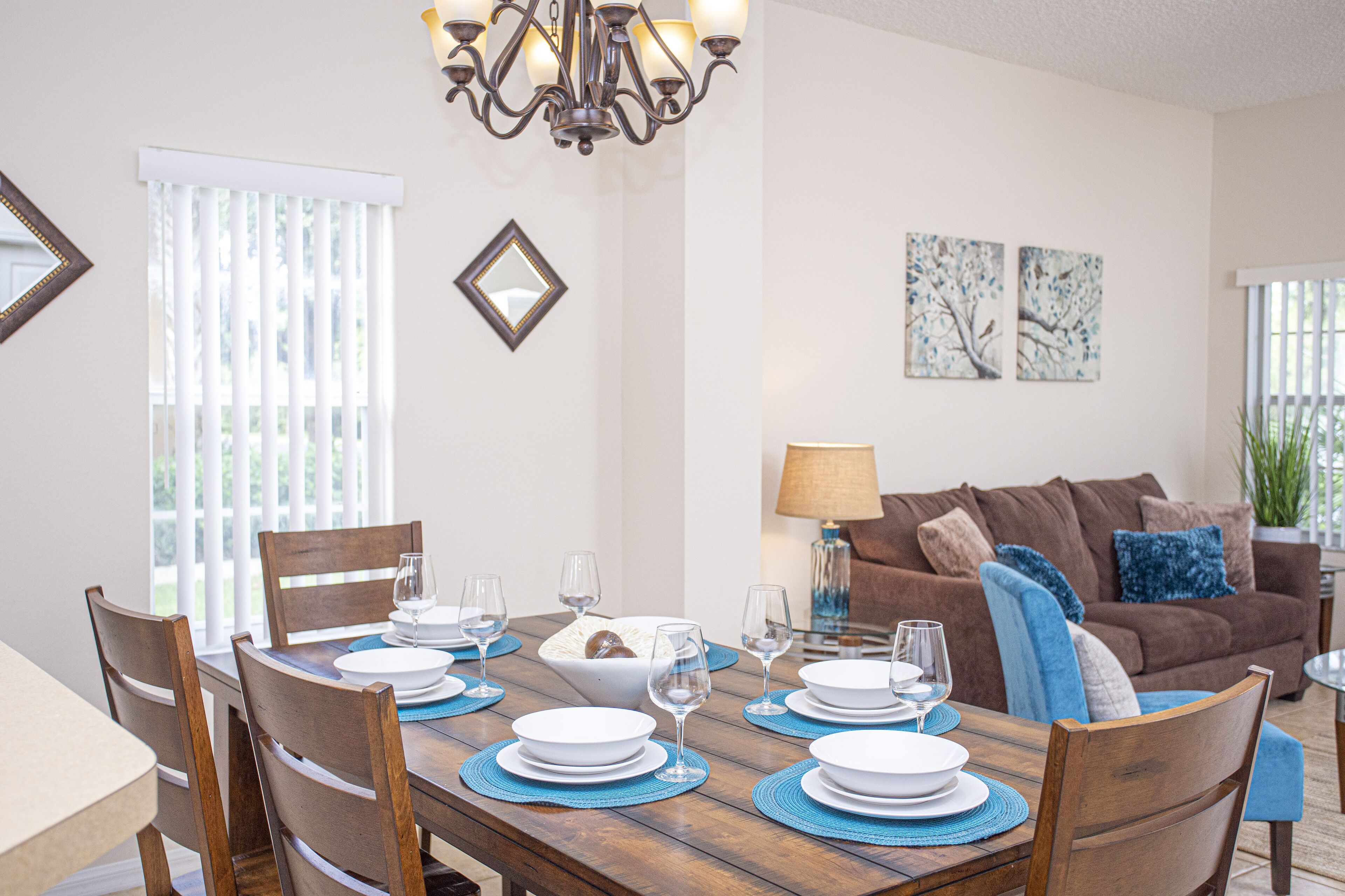 A dining area with a wooden table set for six, surrounded by chairs. There is a chandelier above the table, and a brown sofa with blue cushions in the background.