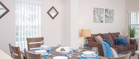A dining area with a wooden table set for six, surrounded by chairs. There is a chandelier above the table, and a brown sofa with blue cushions in the background.