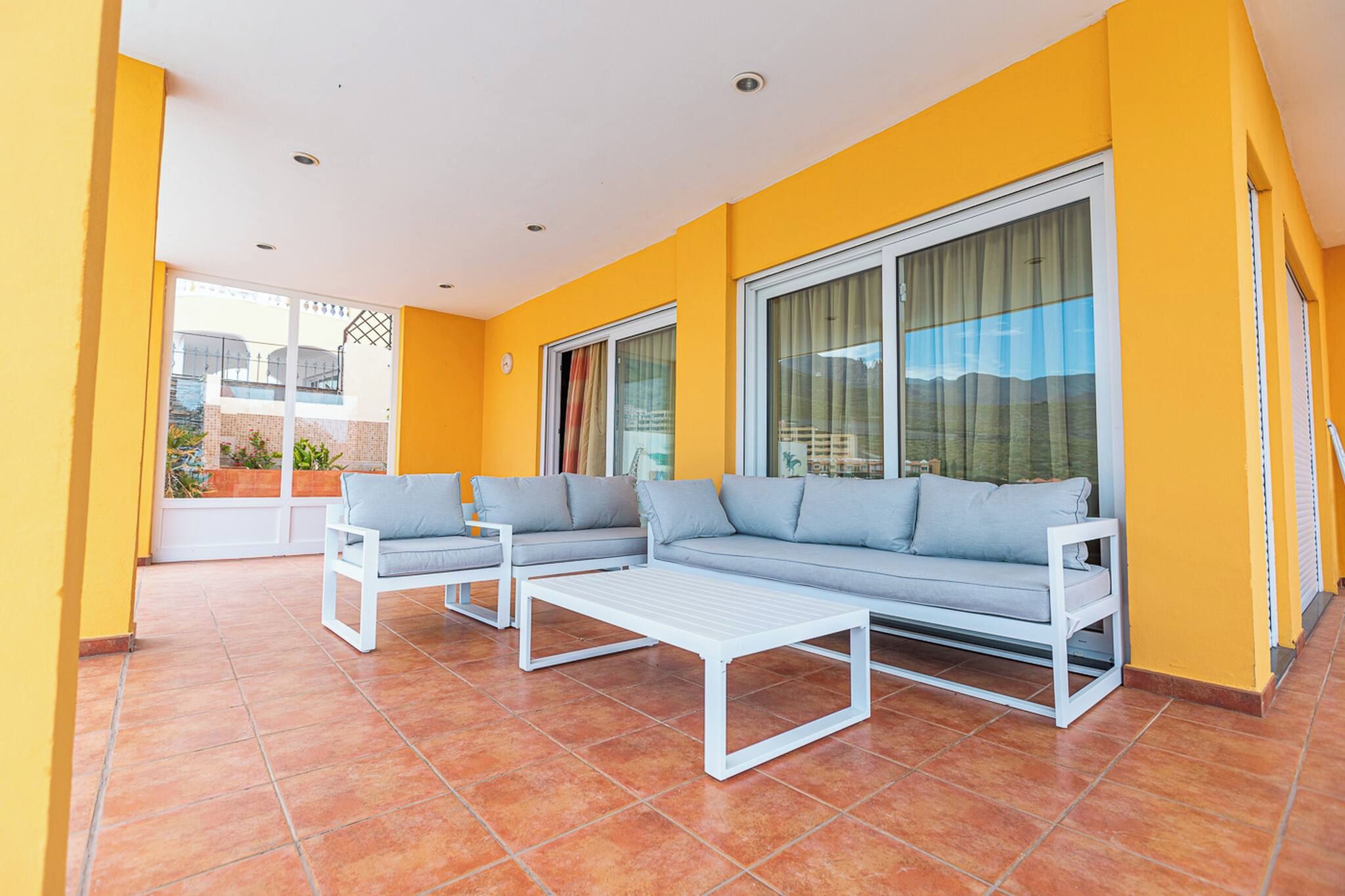 A sunlit patio with a white sofa set, a white coffee table, and a large window with curtains.