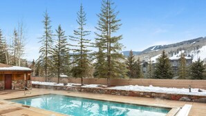 A pool area with a stone wall, a wooden cabin, and a view of mountains and buildings in the background. - Walk to Gondola! - Lionshead - Outdoor Pool & Hot Tubs, Balcony + Private W/D (Vail)