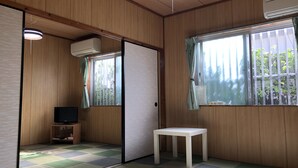 A room with wooden walls, a tatami mat floor, a white table, a TV, and a window with a curtain. - Housei (Zamami)
