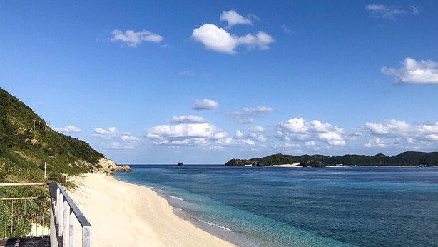 A beach with clear blue water, sandy shore, and distant green hills under a partly cloudy sky. - Housei (Zamami)