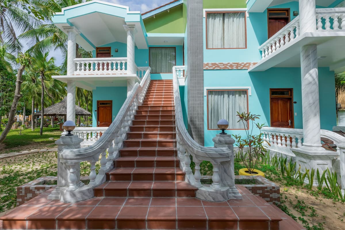 A multi-story building with a staircase leading to a balcony, surrounded by greenery and palm trees.