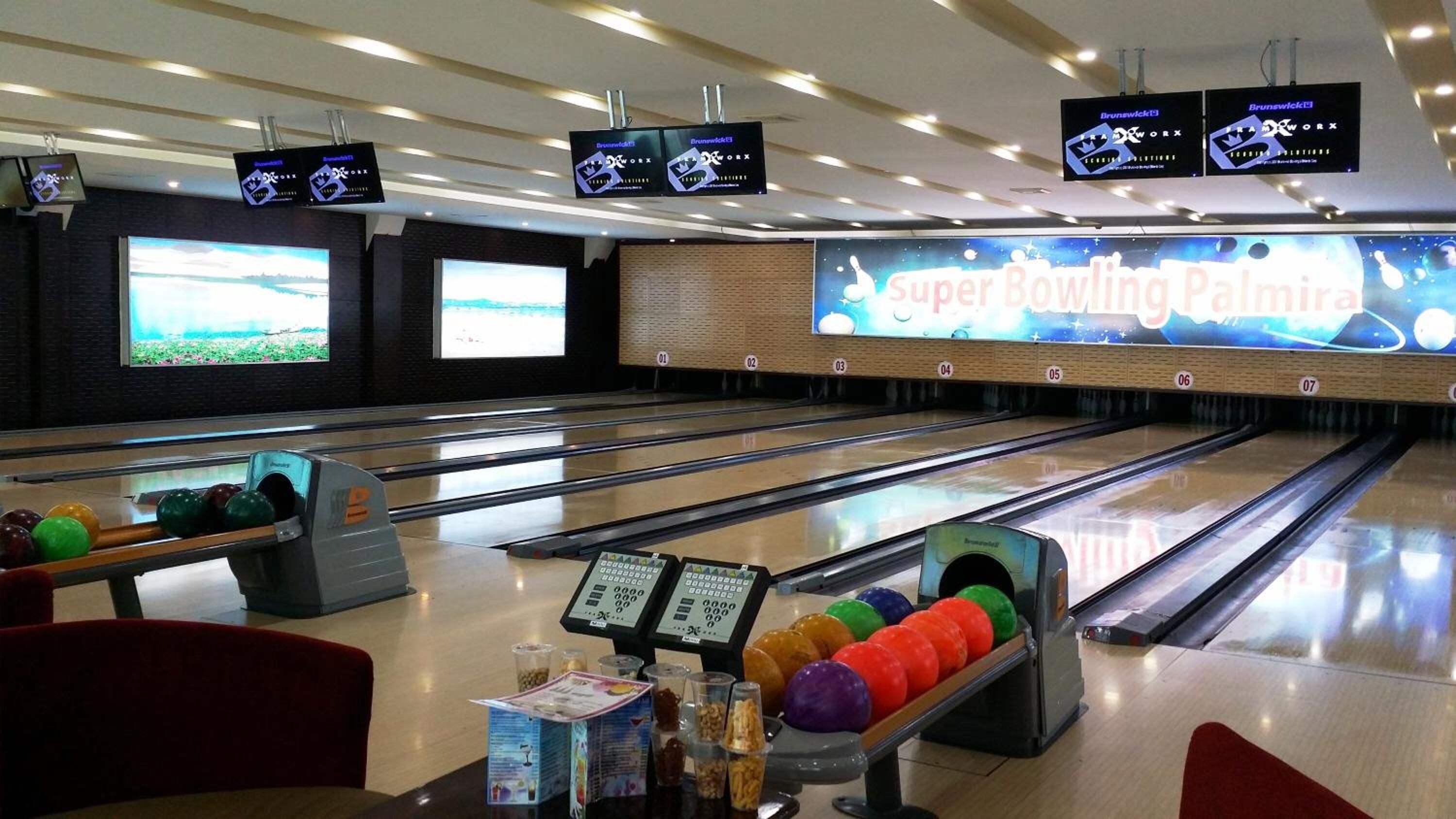 A bowling alley with multiple lanes, a large screen displaying 'Super Bowling Paimira', and several bowling balls on the lanes.