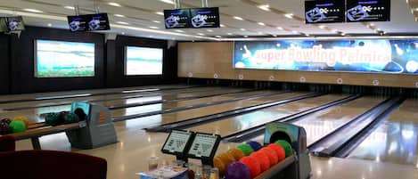 A bowling alley with multiple lanes, a large screen displaying 'Super Bowling Paimira', and several bowling balls on the lanes.