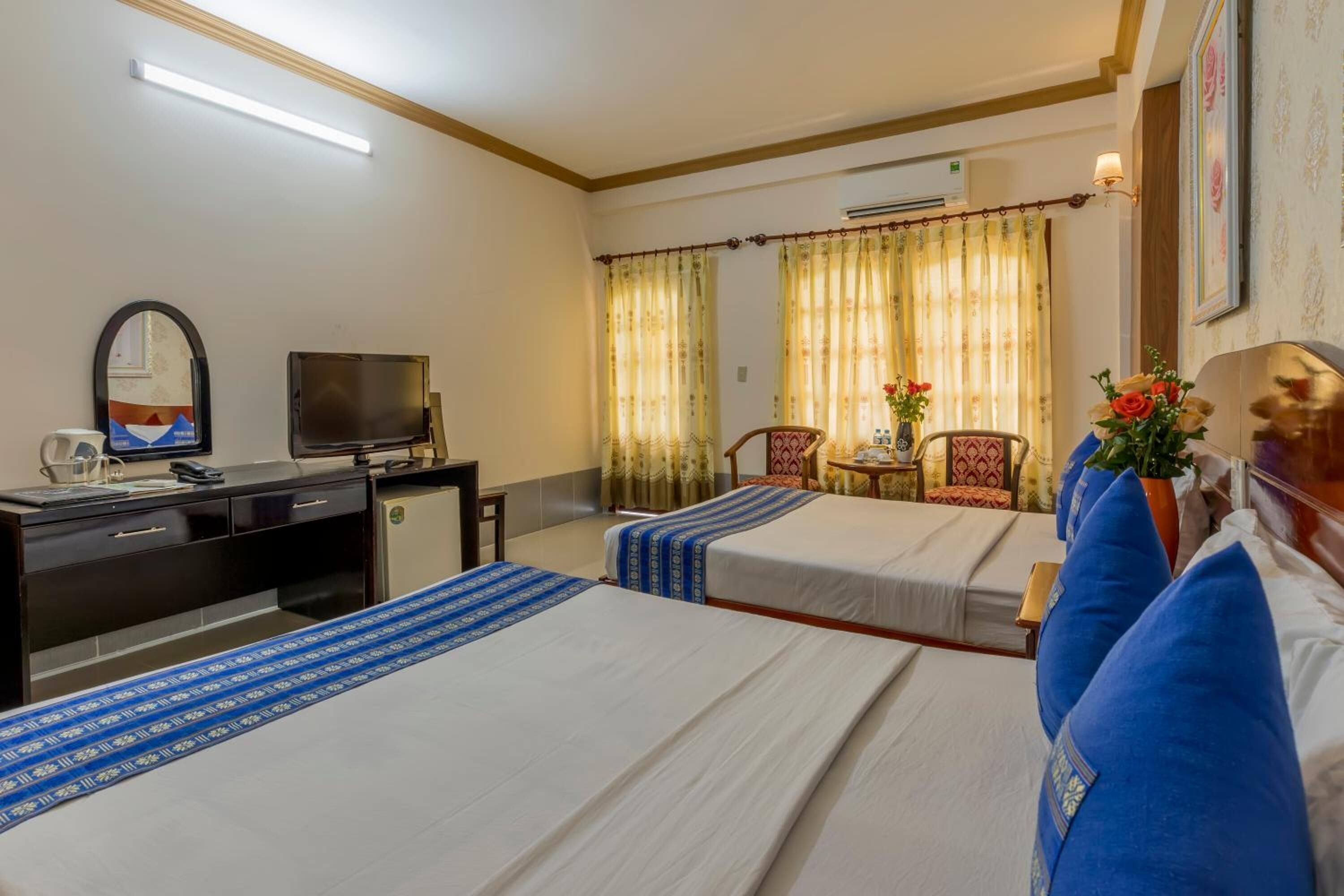Ein Hotelzimmer mit zwei Betten, einem Schreibtisch mit Fernseher, einem Sessel und einem kleinen Tisch mit Blumen.