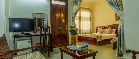 A hotel room with a bed, a desk with a computer, a chair, a table with a vase of flowers, and a window with curtains.