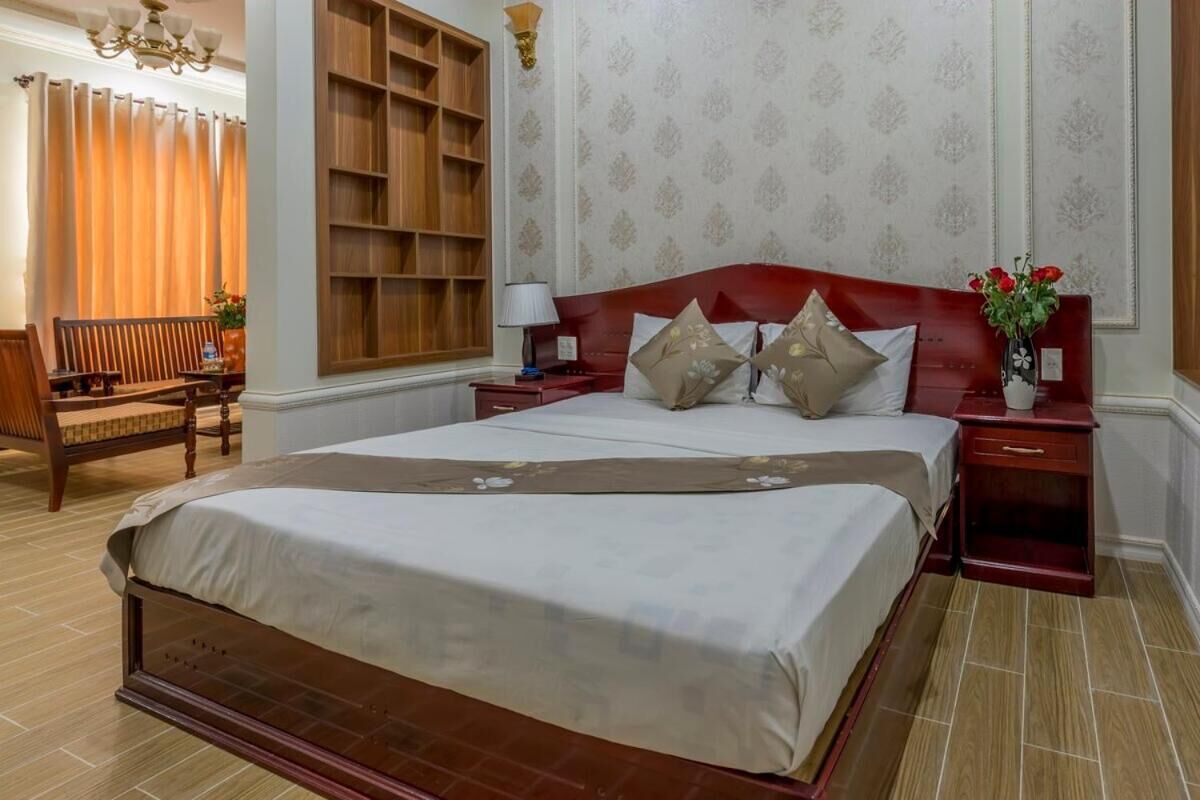 A hotel room with a large bed, wooden headboard, a nightstand, a bedside table, a chair, a desk, and a bookshelf.