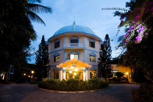 Un edificio grande de varios pisos con techo en forma de cúpula, llamado "Palmira Resort". Cuenta con un balcón central con barandillas, múltiples ventanas y está rodeado de exuberante vegetación y árboles.