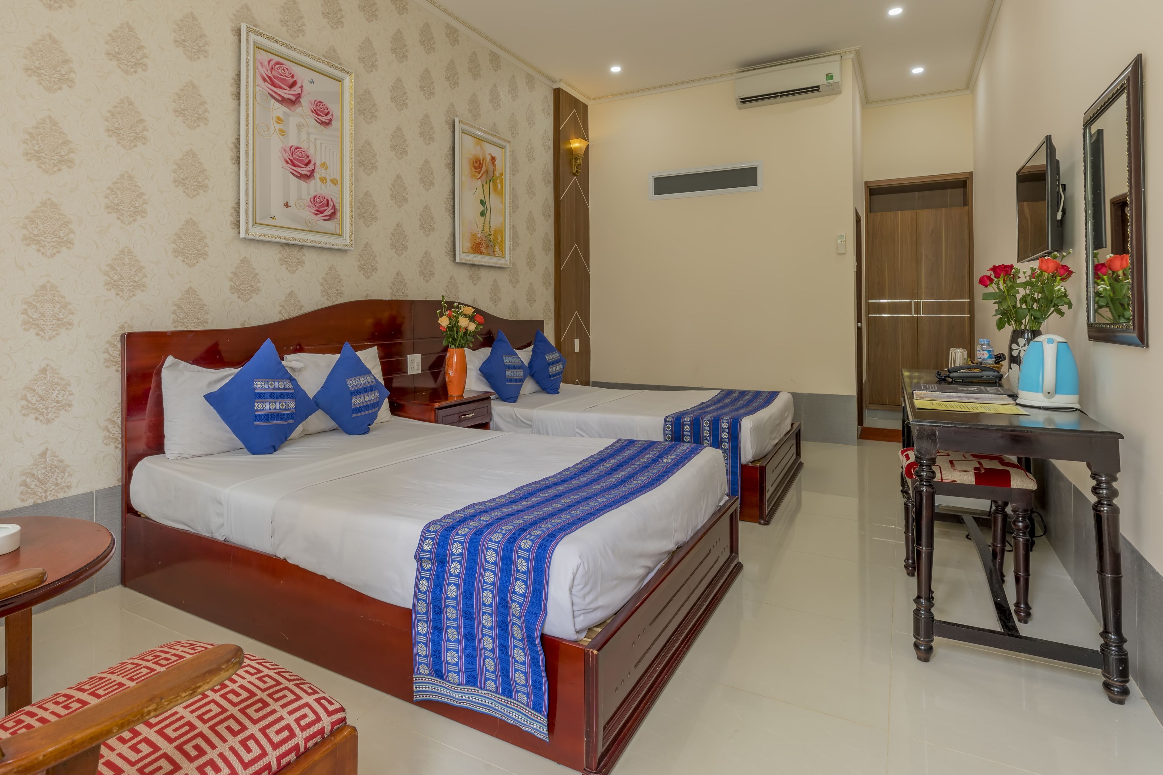 A hotel room with two beds, a wooden headboard, a bedside table with a vase of flowers, and a small table with a chair.