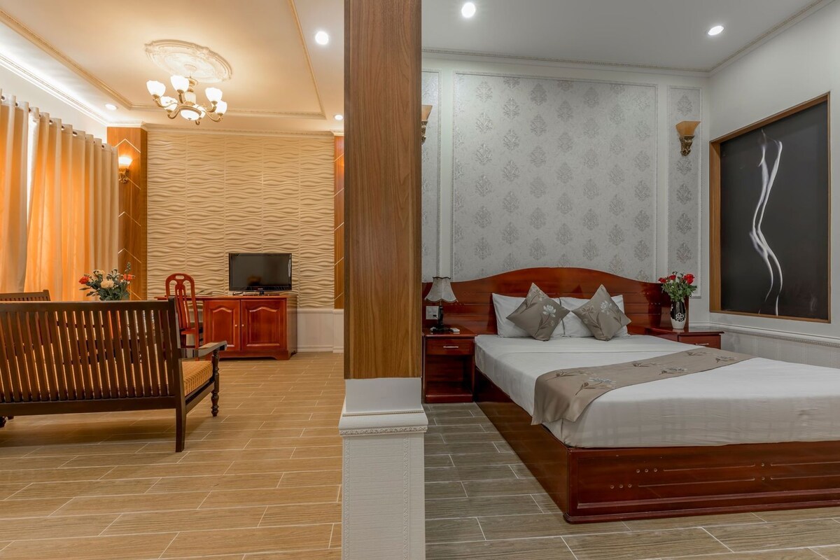 A hotel room with a wooden bed, a TV, a wooden cabinet, a sofa, and a large window.