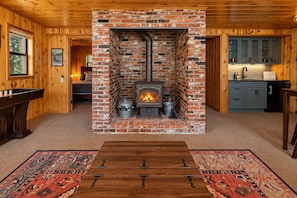 A cozy room with a brick fireplace, wooden walls, a dining table, and a kitchen area.