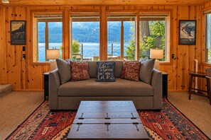 A cozy room with a sofa, a coffee table, and large windows offering a view of a lake and mountains. - Lakefront | Dock | Hot Tub | Deck | Dogs | Views (Leavenworth)