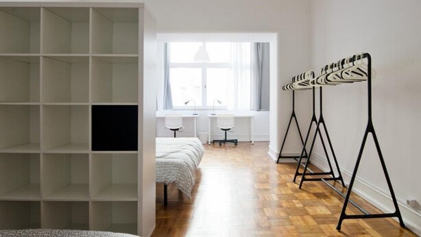 Ein Zimmer mit einer großen, offenen Regaleinrichtung, einem Bett mit gemusterten Bettbezügen, einem Schreibtisch mit zwei Stühlen und einem Fenster mit Vorhängen.