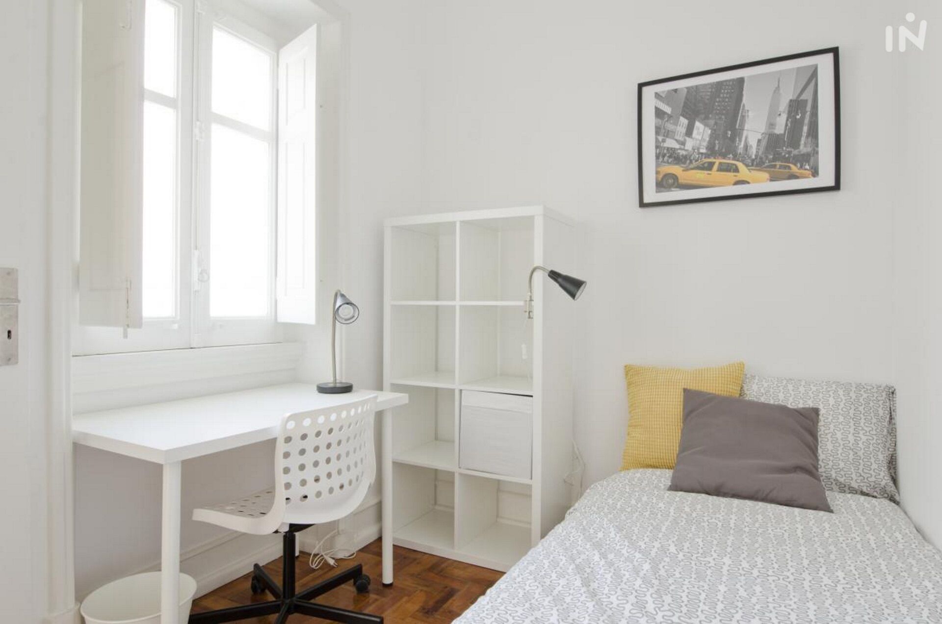 Un dormitorio con un escritorio blanco, una silla, una cama con ropa de cama estampada, una lámpara de noche y un cuadro en la pared.