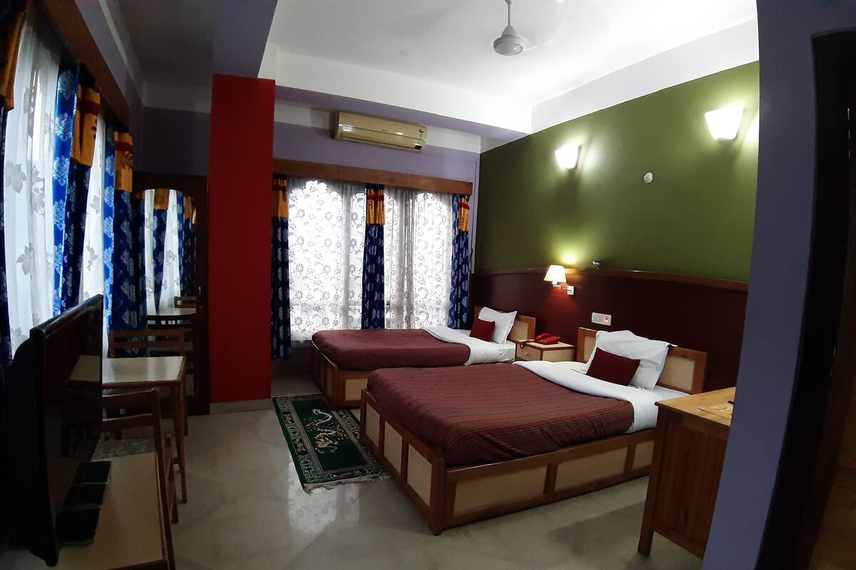 A hotel room with two single beds, a desk with a chair, a TV, and a green carpet.