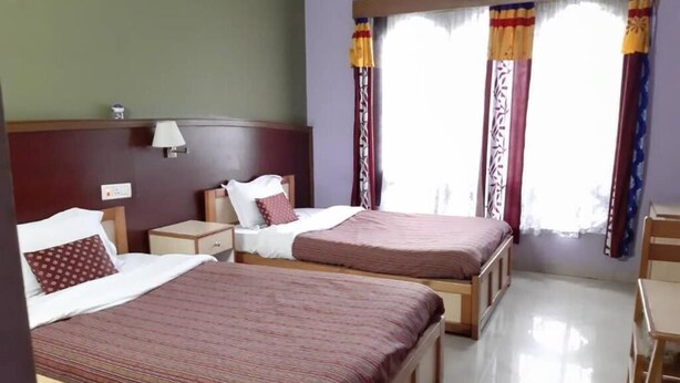 A hotel room with two beds, a nightstand, a lamp, and a window with curtains. - Centennial Hotel 2008 (Phuntsholing)