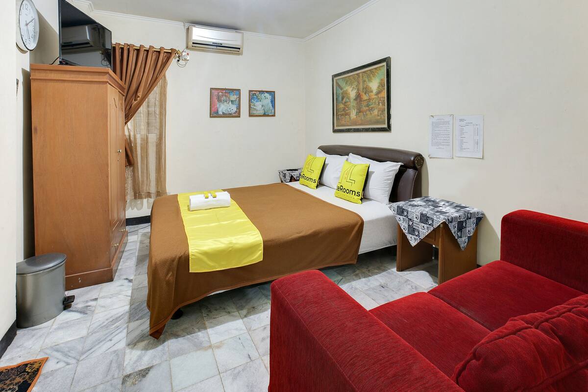 A hotel room with a bed, a red sofa, a wooden wardrobe, a trash bin, and a painting on the wall.
