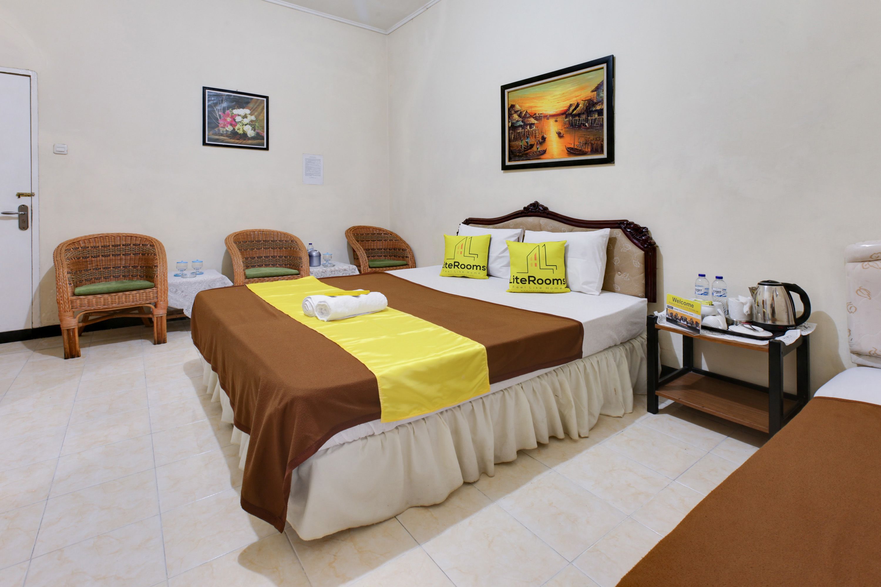 A hotel room with a bed, two wicker chairs, a bedside table with a kettle, and a painting on the wall.