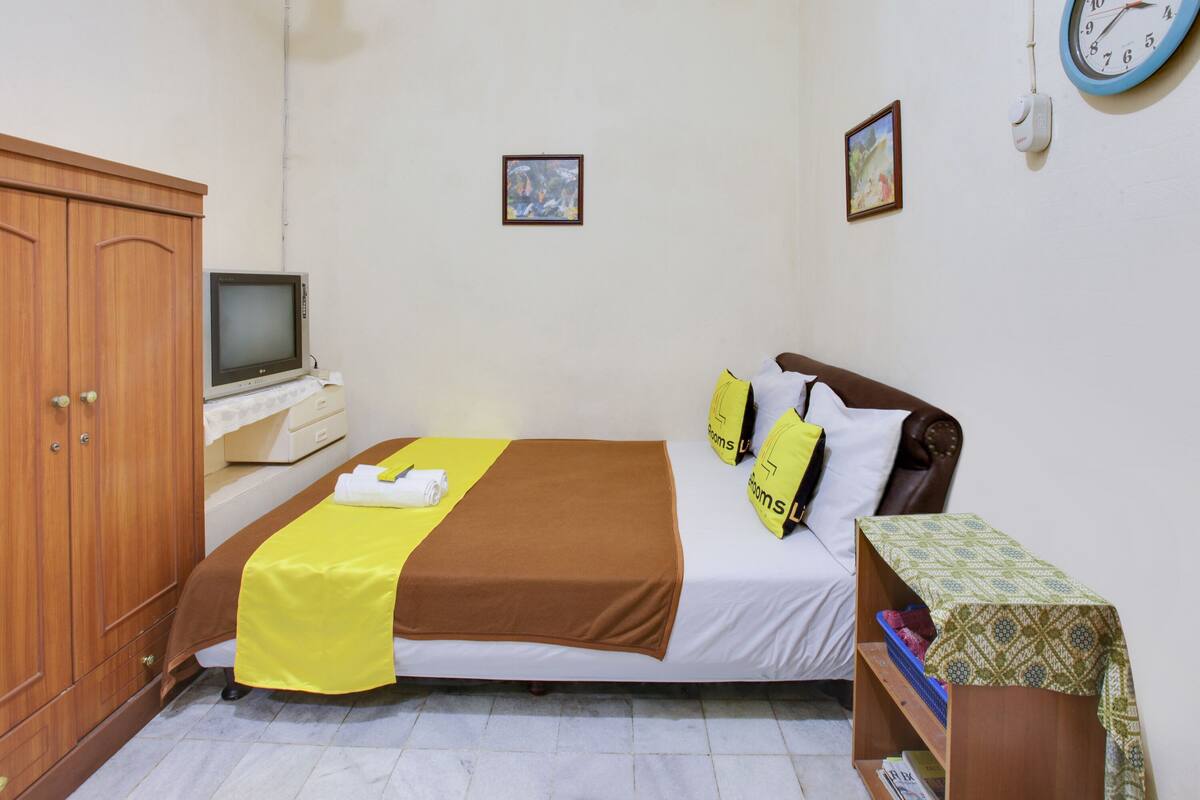 A hotel room with a bed, a wooden wardrobe, a television, and a small table with a patterned tablecloth.