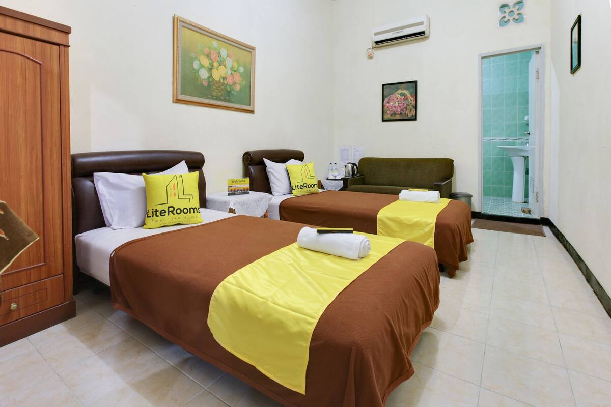 A hotel room with two beds, each with a brown bedspread and a yellow pillow. There is a wooden wardrobe, a painting on the wall, and a bathroom visible through an open door.